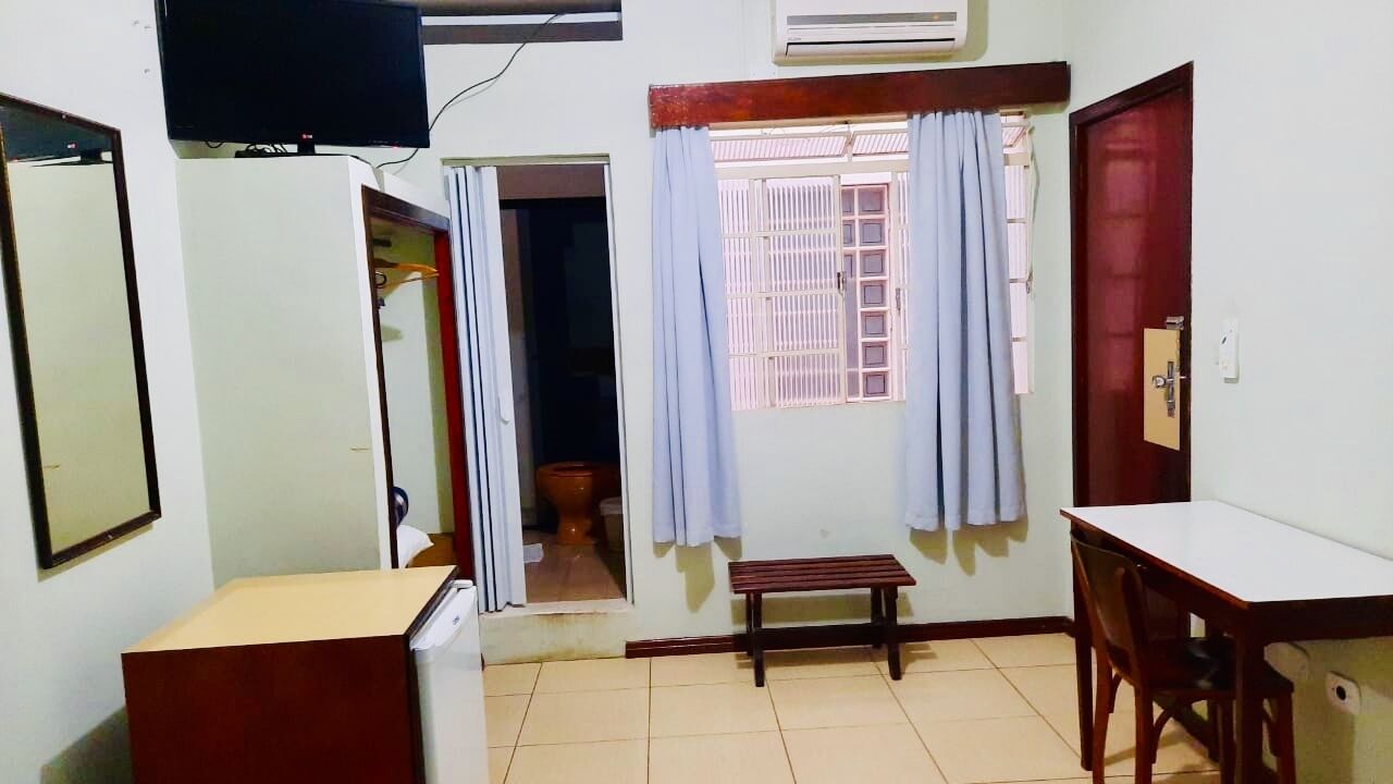 Una habitación con escritorio, banca, armario, televisor y ventana con cortinas.