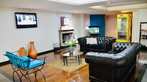 Un salon avec un canapé en cuir noir, un fauteuil bleu, une petite table sur laquelle est posé un vase, et un téléviseur fixé au mur.