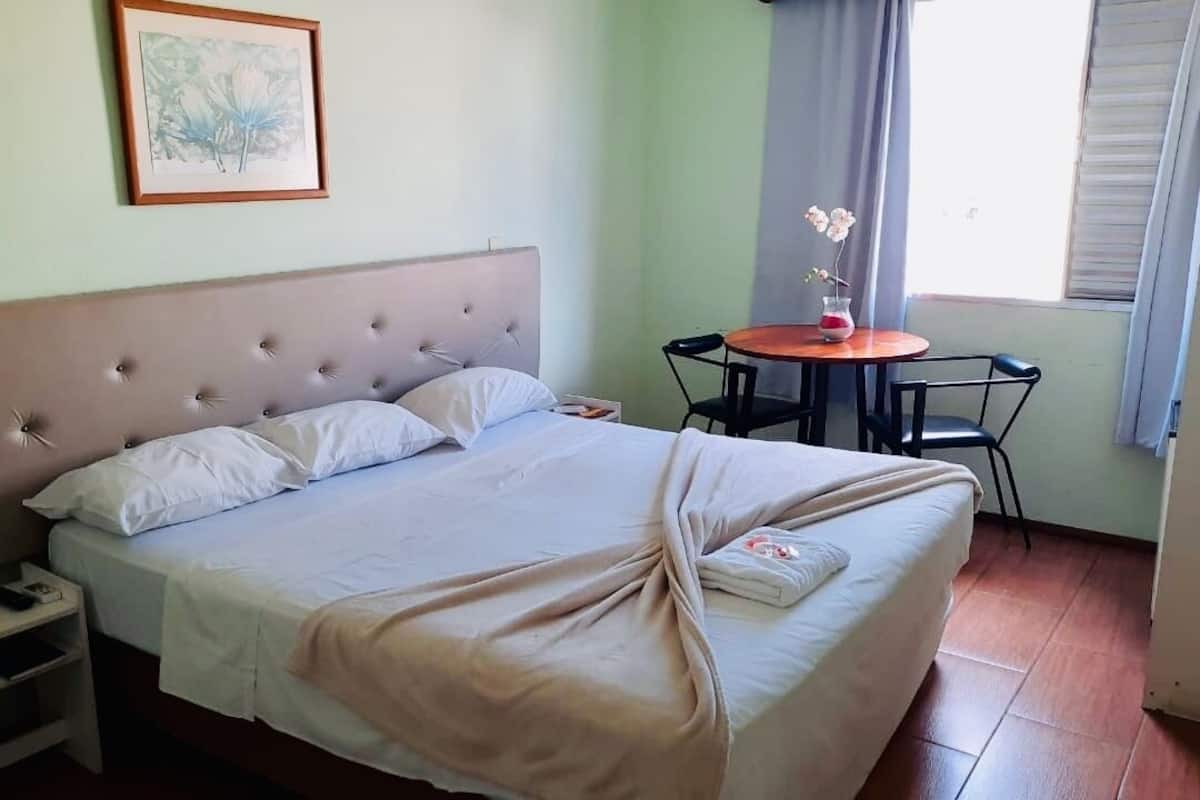 Un llit ben fet amb un capçal, una taula de nit amb un telèfon, una taula petita amb un ram de flors i una finestra amb cortines.