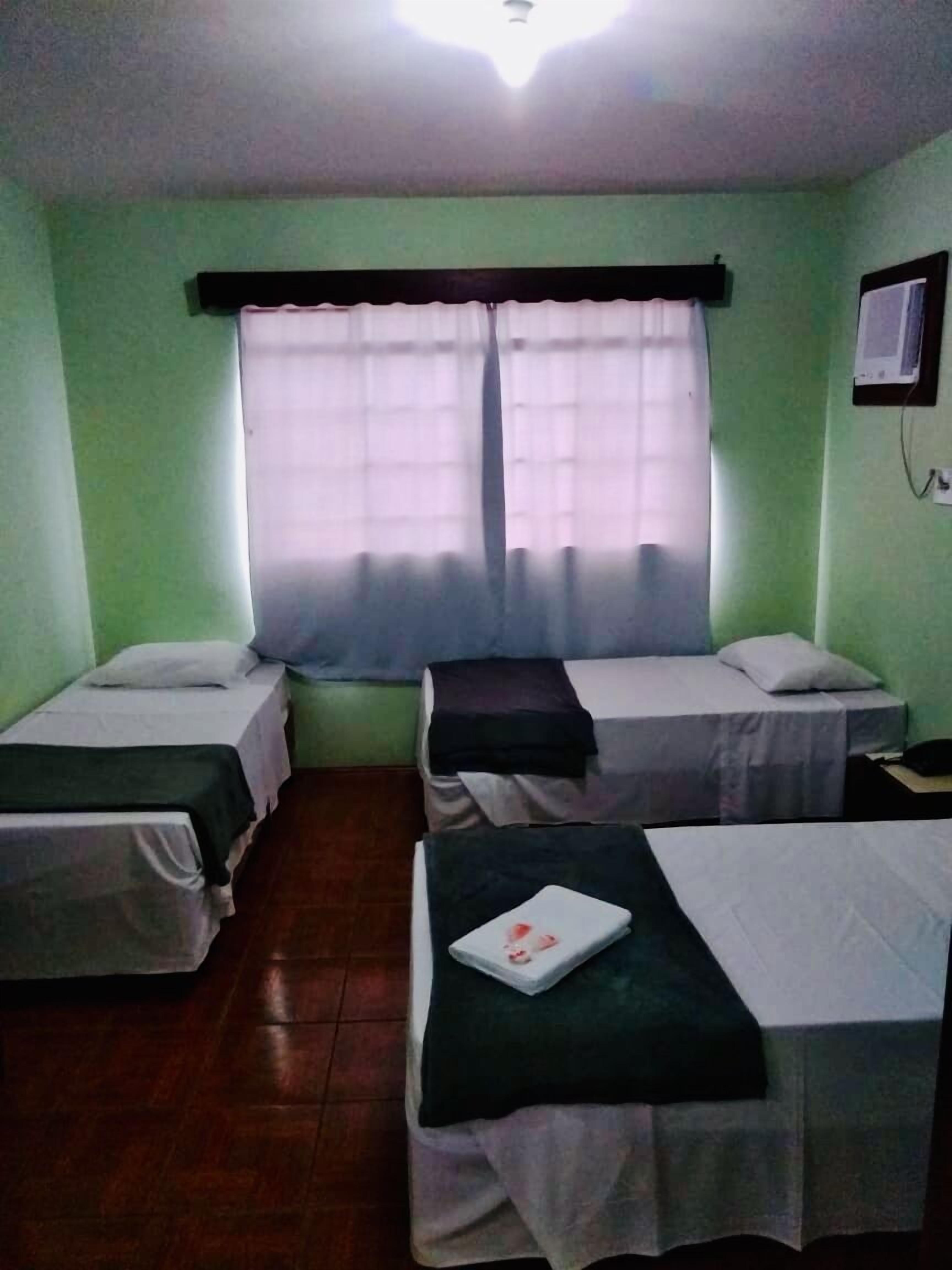 A room with two single beds, a window with sheer curtains, a wall-mounted TV, and a tiled floor.