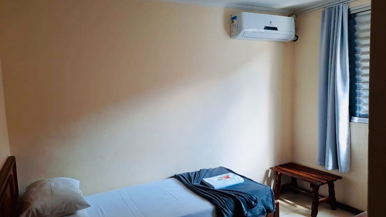 A small, simple room with a bed, a bedside table, a window with curtains, and an air conditioning unit.