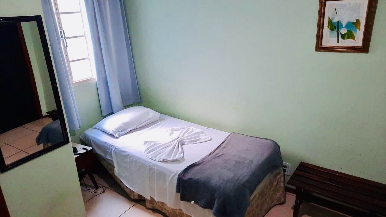 A small, single-bed room with a bed, a desk, and a chair. There is a window with curtains, a framed picture on the wall, and a mirror on the wall.