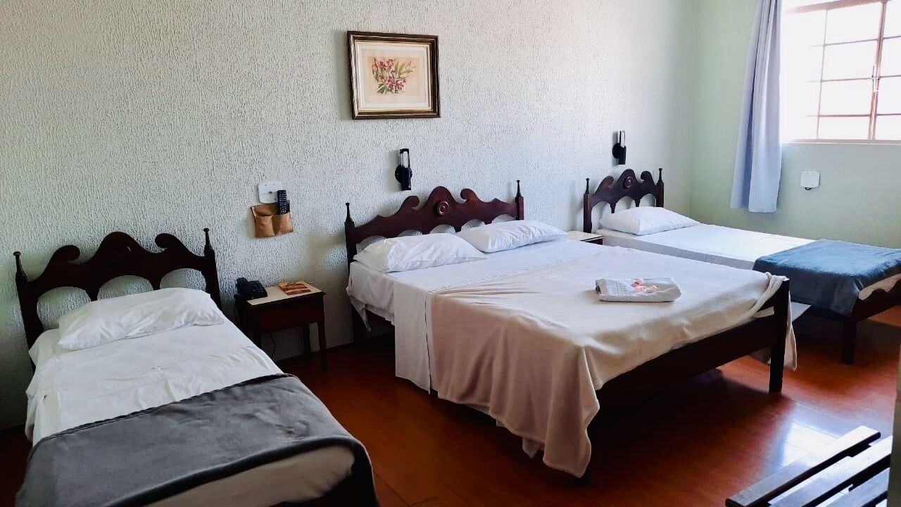Une chambre d’hôtel avec trois lits, une tête de lit en bois, une lampe fixée au mur et une fenêtre avec des rideaux.