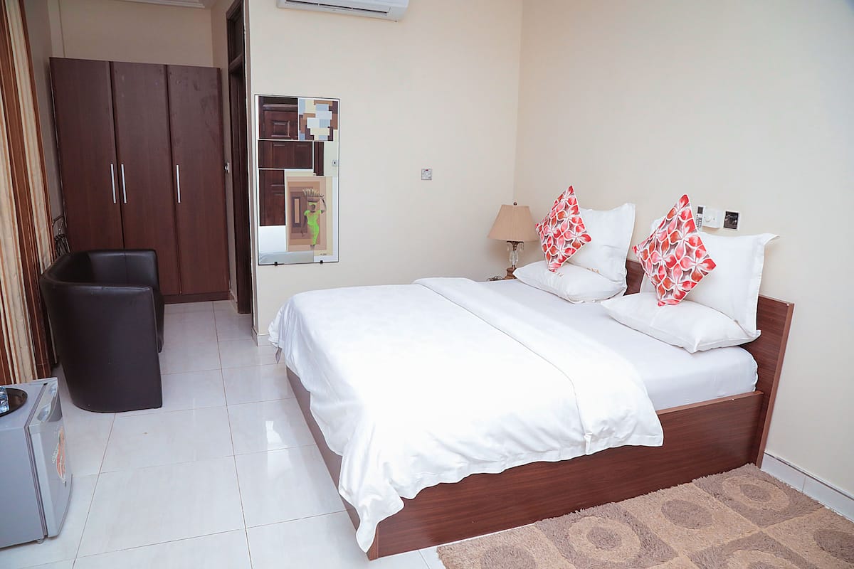 A bedroom with a bed, a wardrobe, a mirror, a lamp, and a trash bin.
