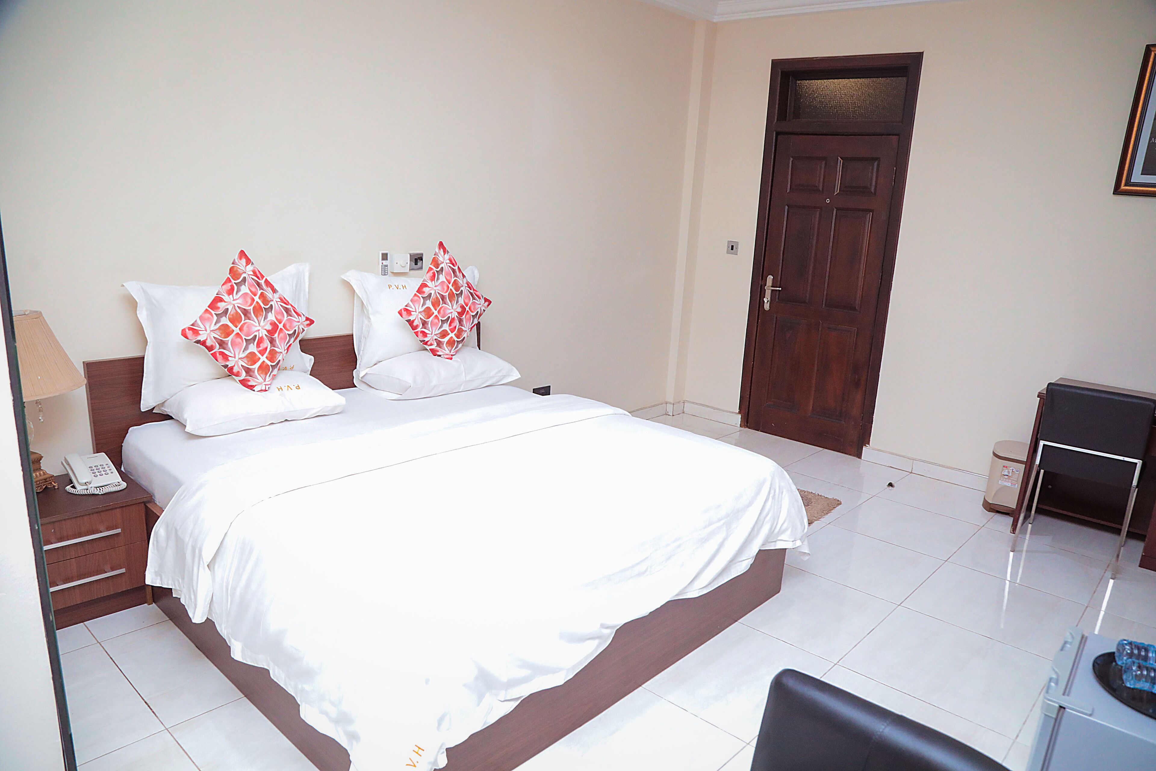 A hotel room with a large bed, two pillows with a red and white pattern, a wooden door, and a desk with a chair.