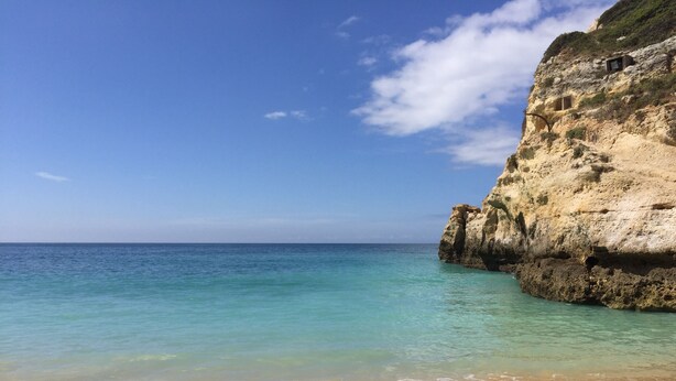 A beach with clear turquoise water, a sandy shore, and a rocky cliff.