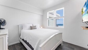 A bedroom with a bed, a window with a view, a fan, and a TV on a stand. - Sandpiper Oceanfront 5F (Jacksonville Beach)