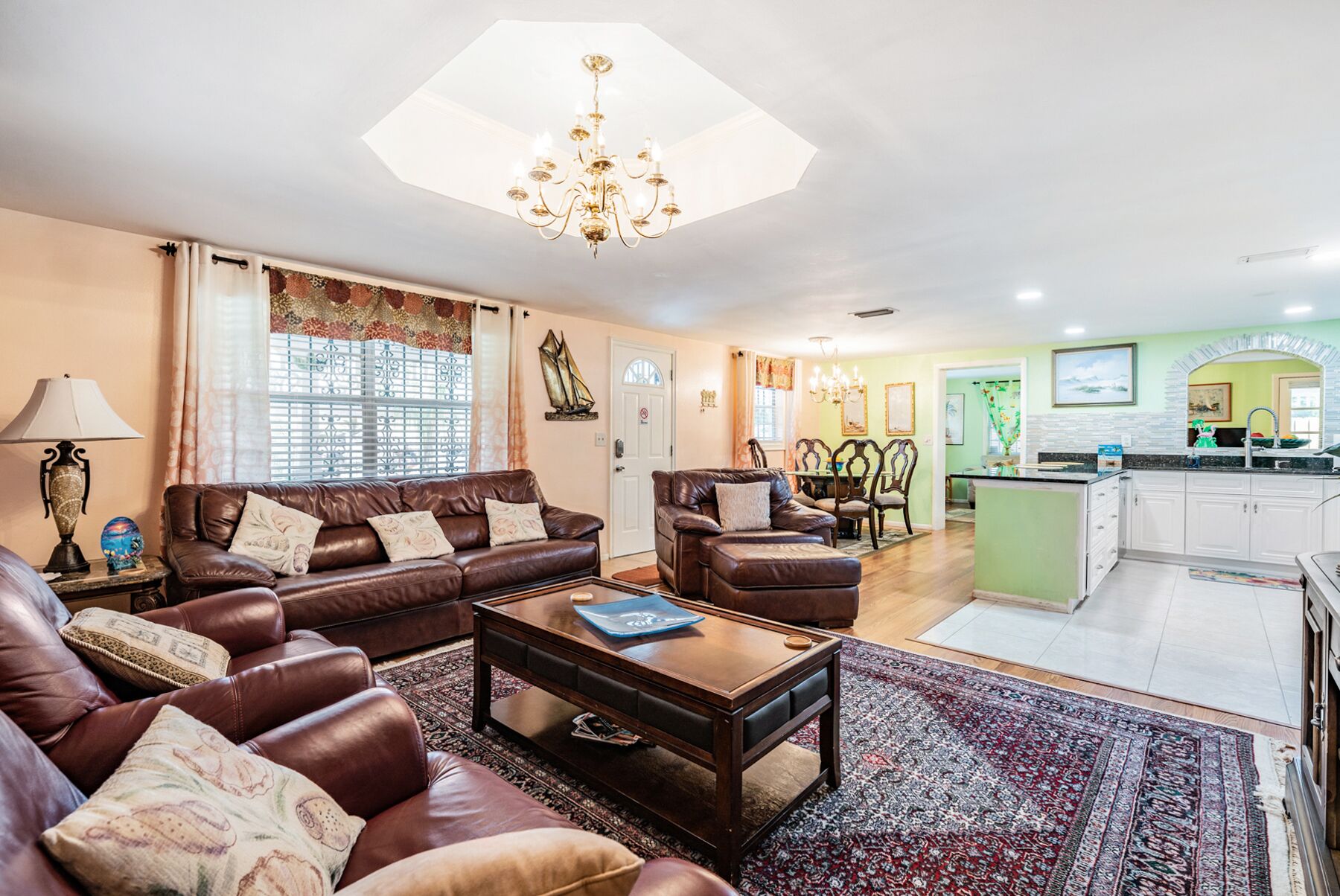 A living room with a brown leather sofa, a wooden coffee table, and a patterned rug. There is a dining area with a table and chairs, and a kitchen with white cabinets and a green backsplash.