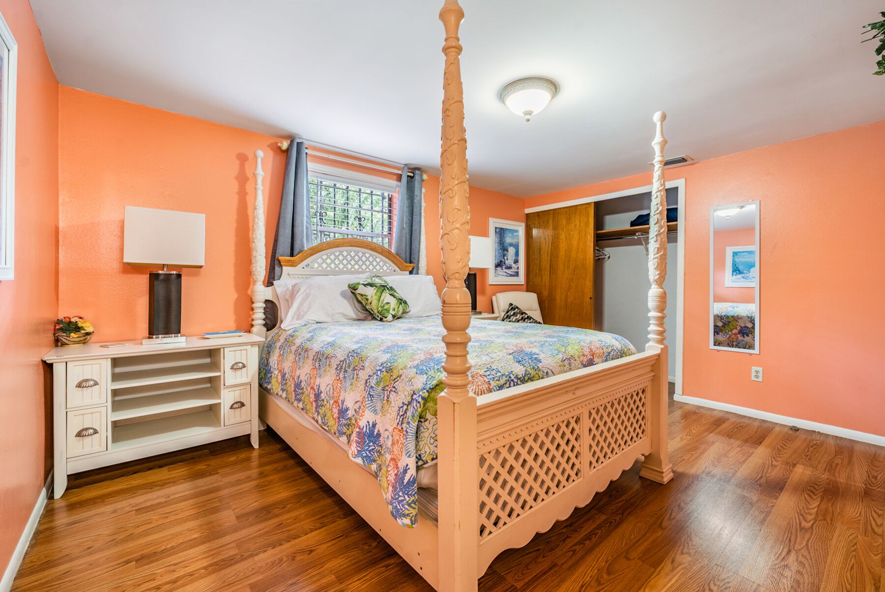 Uma cama com dossel adornada com colcha floral, um criado-mudo com abajur, um aparador e uma janela com cortinas.