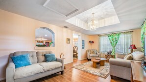 A spacious living room with a grey sofa, a smaller armchair, and a coffee table. There is a chandelier hanging from the ceiling, and a large window with green curtains. - #1 LOCATION  - Walk to Amusement Parks from PERFECT HOME, Great for Families 🏠 (Tampa)
