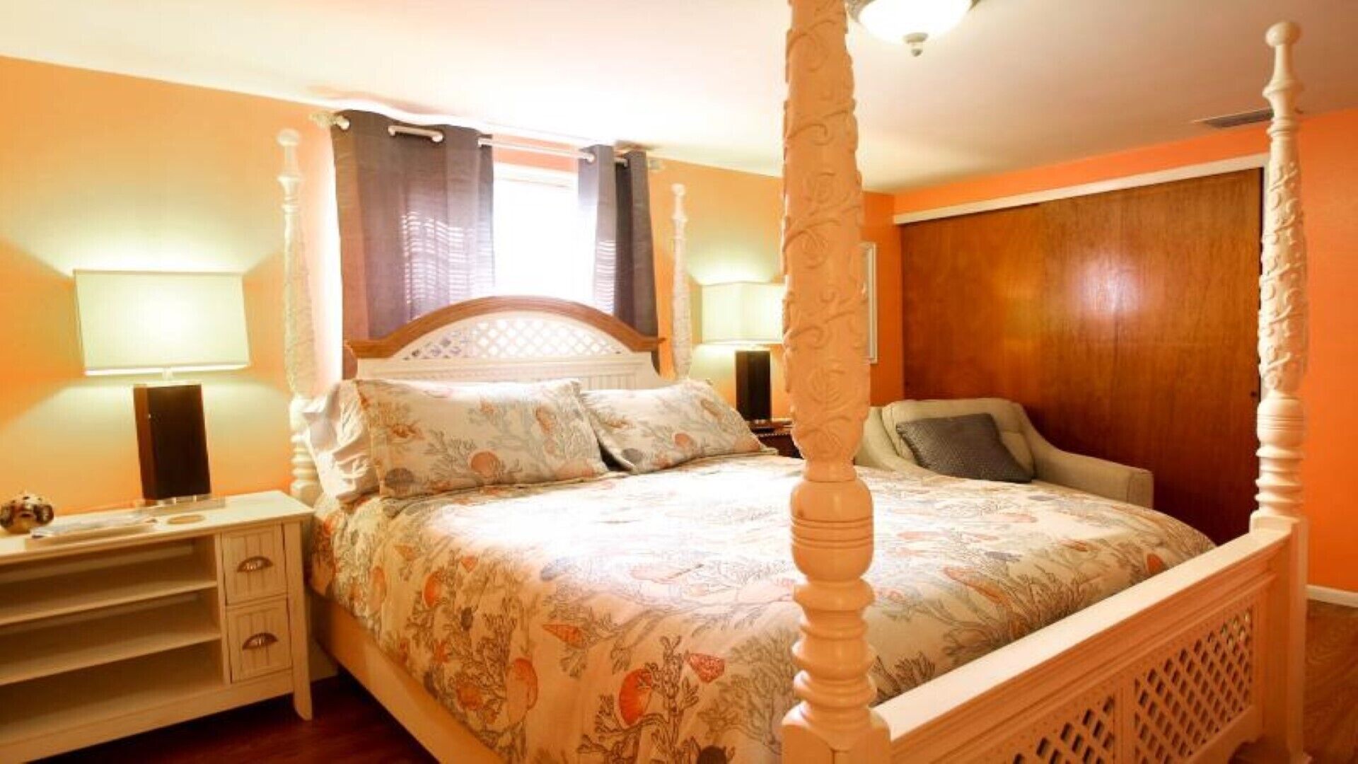Una cama con dosel, ropa de cama floral, una mesita de noche con lámpara y un armario de madera en una habitación con luz cálida.