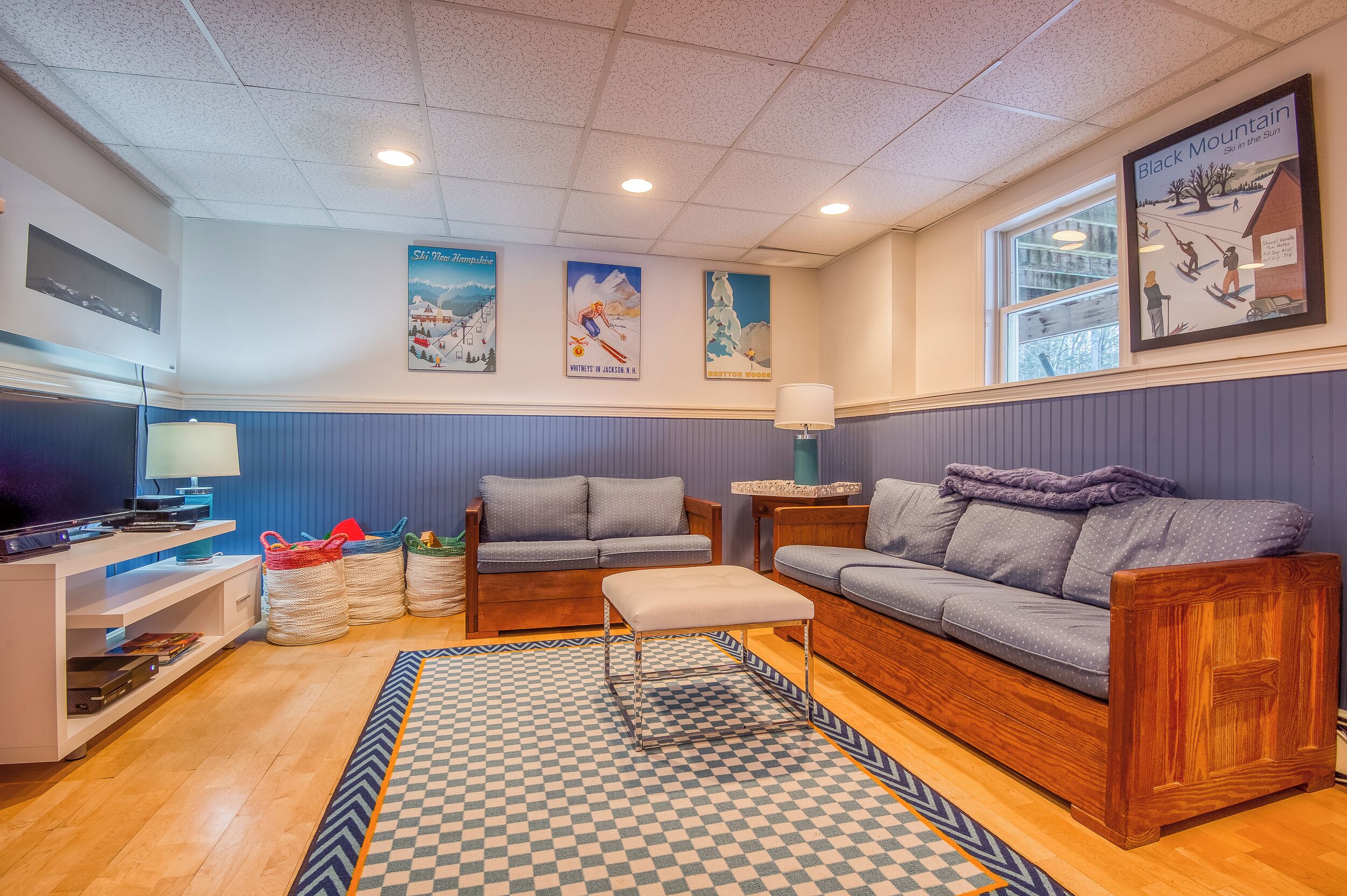 Un salon chaleureux avec un tapis à carreaux, deux canapés, un banc en bois et un téléviseur posé sur un meuble blanc.