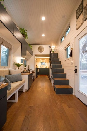 Un couloir étroit doté d’un plancher en bois, d’un escalier, d’un coin cuisine avec un évier et un plan de travail, et d’un espace détente avec un banc et des coussins.