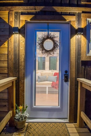 Une porte extérieure en bois avec un panneau de verre, une couronne de fleurs, un banc avec des coussins et une plante en pot.
