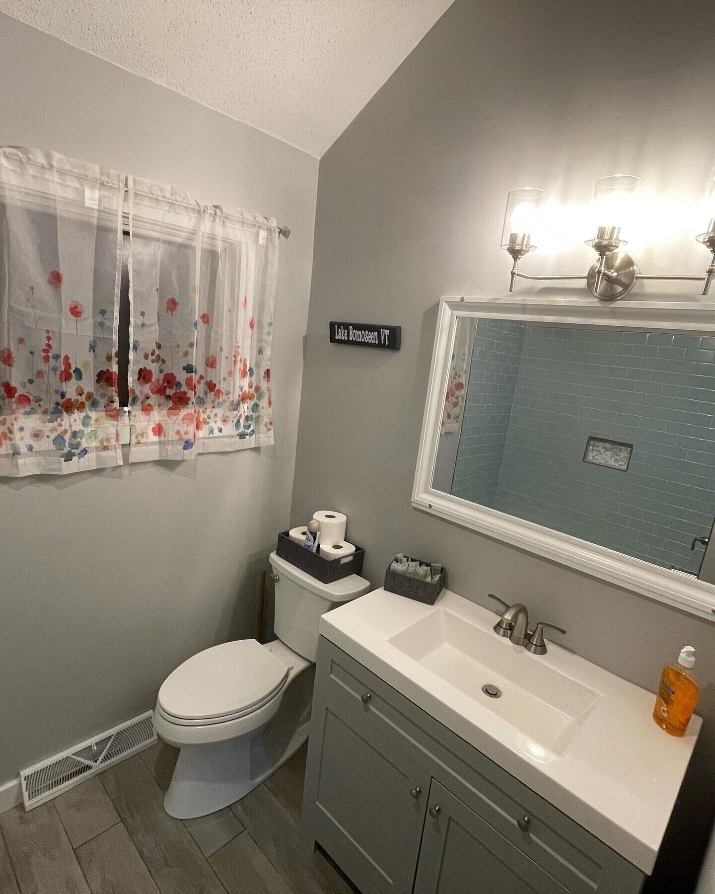 Une salle de bain équipée de toilettes, d’un lavabo et d’un espace douche. Un panneau est affiché sur le mur, sur lequel on peut lire : « Lac Bonosee, Vermont ».