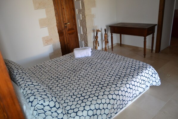 A bedroom with a patterned bedspread, a wooden door, a small wooden table, and a tiled floor.