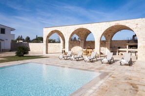 A pool area with a stone wall and arches, surrounded by lounge chairs.