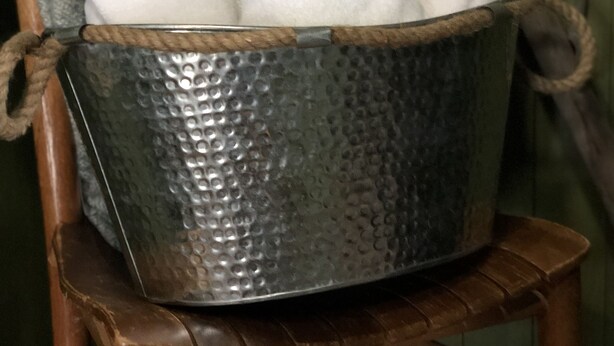 A metal bucket with a textured surface containing several white towels, placed on a wooden chair with a rope handle.