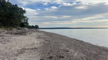 Ein Kiesstrand mit Bäumen links und ruhigem Wasser im Hintergrund.