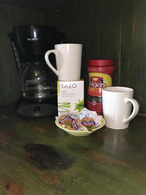 Una cafetera, una caja de té Tazo, café instantáneo Folgers y dos tazas sobre una superficie de madera.