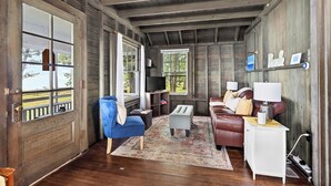 Una habitación rústica con paredes de madera, un ventanal grande, una silla azul, un sofá marrón y un armario blanco.