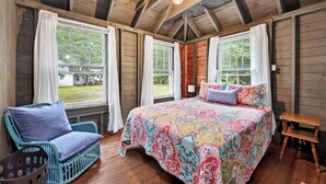 Habitación de cabaña de madera con una cama grande, dos ventanas, una silla y una mesita.