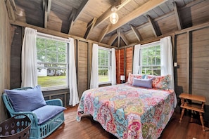 Quarto de cabana de madeira com uma cama grande, duas janelas, uma cadeira e uma mesinha.
