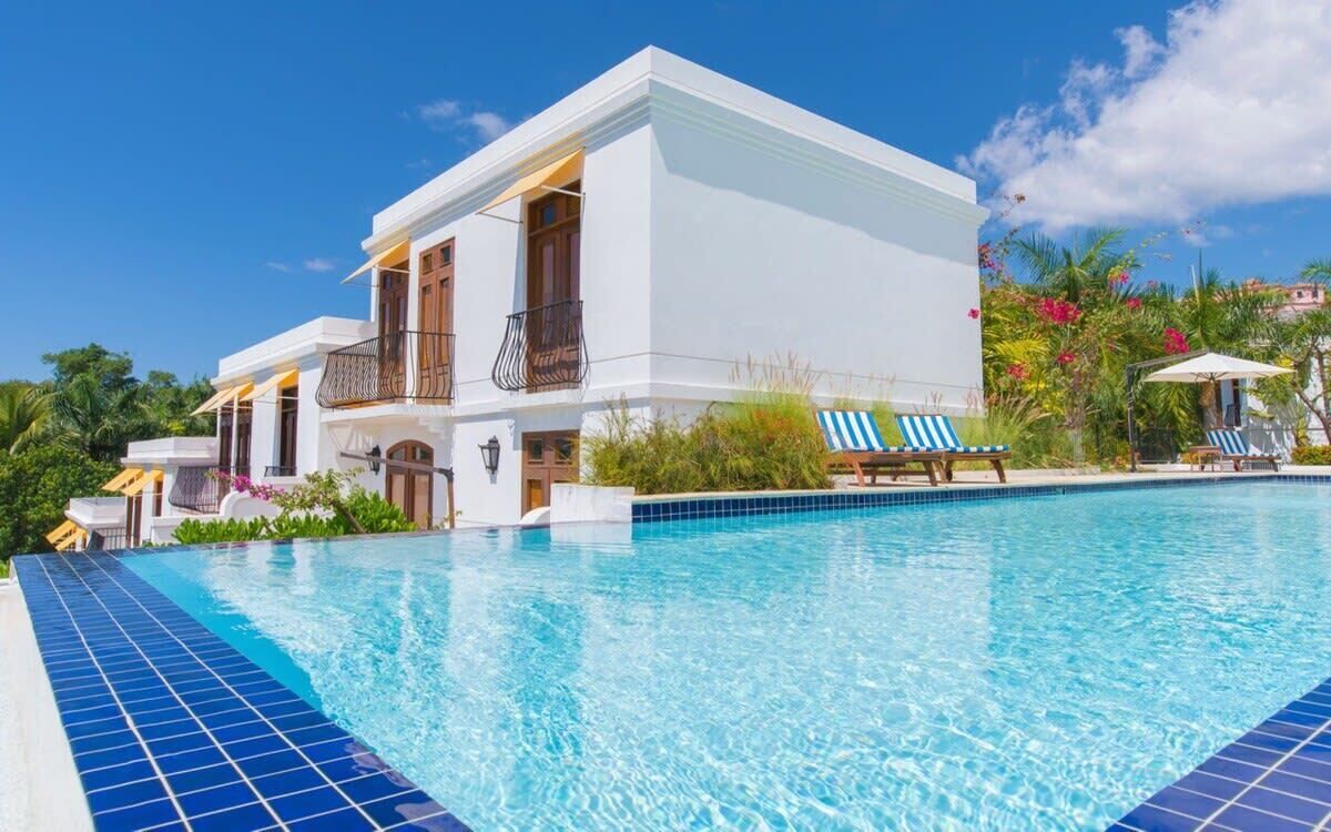 A modern white building with a swimming pool, surrounded by tropical plants and a clear blue sky.