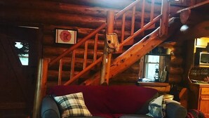 L’intérieur d’un chalet confortable, avec des murs en bois, un escalier en bois, un canapé bleu agrémenté de coussins, un tapis à motifs et un téléviseur posé sur un meuble en bois.