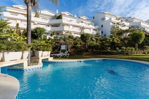 A swimming pool with lounge chairs, surrounded by lush greenery and white buildings.