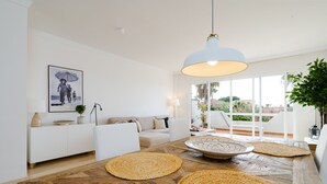 A modern living room with a wooden coffee table, white sofas, a hanging light fixture, and a large window with a view of greenery.