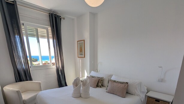 A bedroom with a large bed, a chair, a bedside table, and a view of the sea through a window. - 201 Apartamento La Duna (Tarifa)