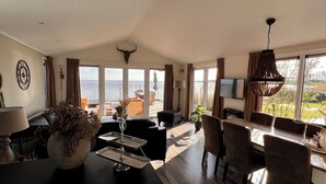 Ett rymligt vardagsrum med ett stort matbord, bekväma sittplatser, en TV och utsikt över havet genom stora fönster.
