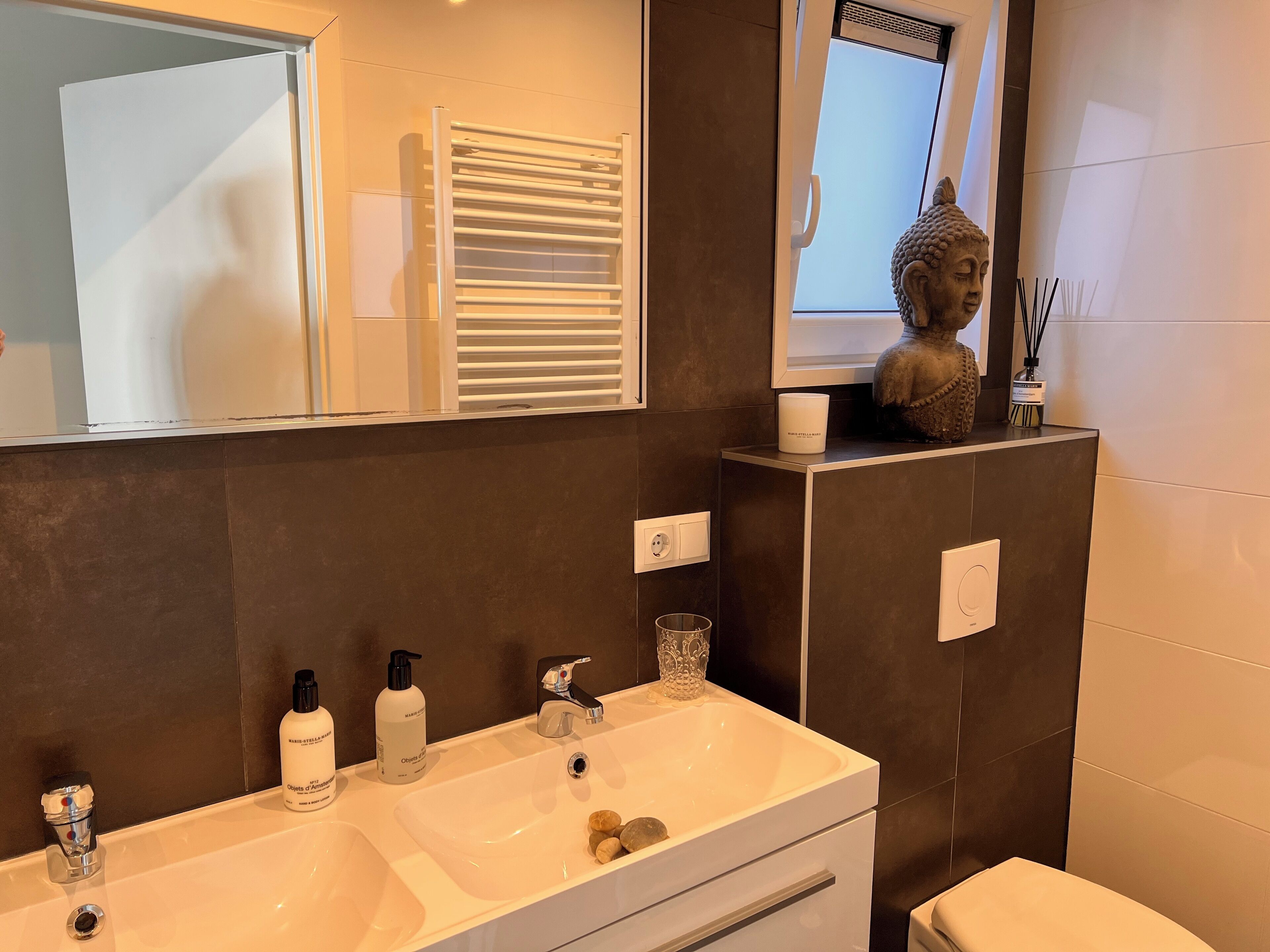 A modern bathroom with a dual sink vanity, a large mirror, a towel rack, a Buddha statue, and a small shelf with a candle and reed diffuser.