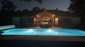 A swimming pool with a wooden deck in front, a covered patio area with outdoor furniture, and a house with a gabled roof in the background. - Villa Esprit de Famille in Moliets et Maa, close to the ocean and golf course (moliets et maa)