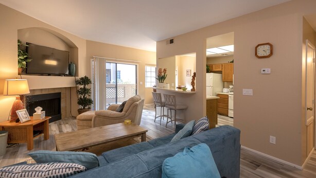 A living room with a sofa, a TV mounted on the wall, a fireplace, and a kitchen area visible through an open door.
