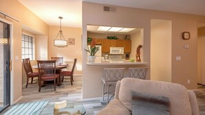 A living room with a beige sofa, a glass coffee table, and a dining area with wooden chairs and a small island with a microwave.