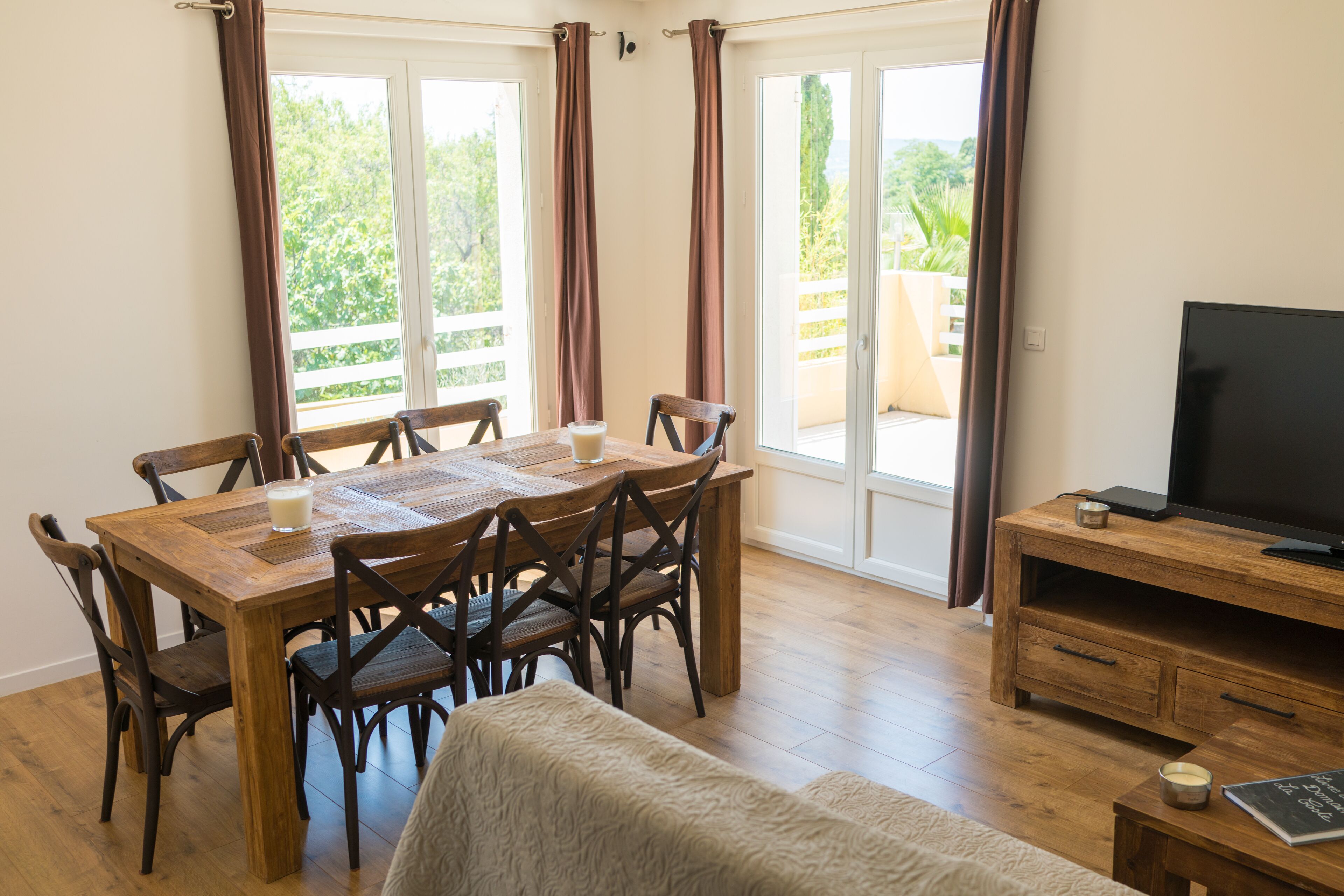 A dining area with a wooden table and chairs, a television on a wooden stand, and a view of greenery through the windows.