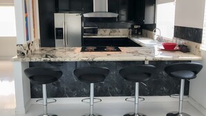 A modern kitchen with black cabinets, a marble countertop, a stainless steel refrigerator, a microwave, and a gas stove. There are three black bar stools at the island. - Just listed - The Enticing Villa Cardona, Magnesia Falls, Rancho Mirage (Rancho Mirage)