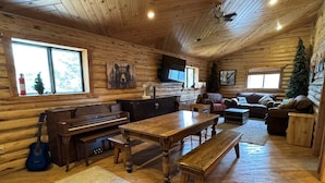 A log cabin interior with a wooden dining table, benches, a piano, a fireplace, and a large window.
