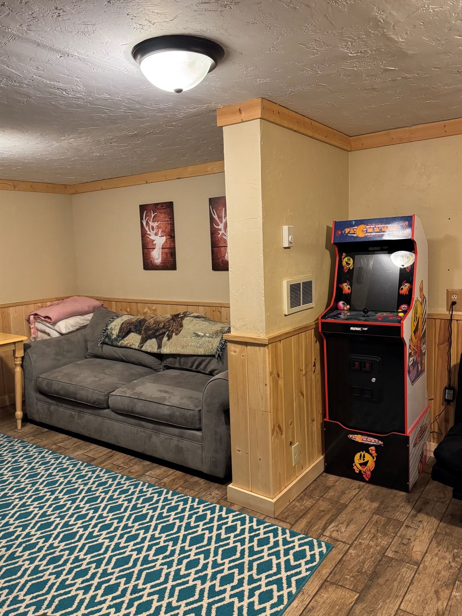 Una habitación con un sofá gris, un tapete estampado, un armario de madera y una máquina arcade de Pac-Man.