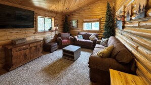 A log cabin interior with a large flat-screen TV, multiple seating areas, and a fireplace.