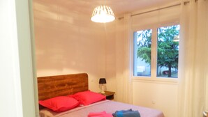 A bedroom with a wooden headboard, a bed with pink and blue linens, a bedside lamp, and a window with a view of trees.