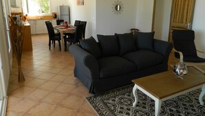 A living room with a black sofa, a wooden coffee table, and a patterned rug. There is a dining area with a table and chairs, and a kitchen in the background.