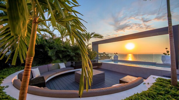 Un moderno espacio lounge al aire libre con una distribución de asientos curva, una piscina y vistas al atardecer sobre el agua.