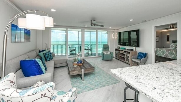 A spacious living area with a large window overlooking the ocean, a kitchen area with a dining table, and a seating area with a sofa and armchairs. - Crystal Sands Tower Unit 903 - Beachfront (Sarasota)