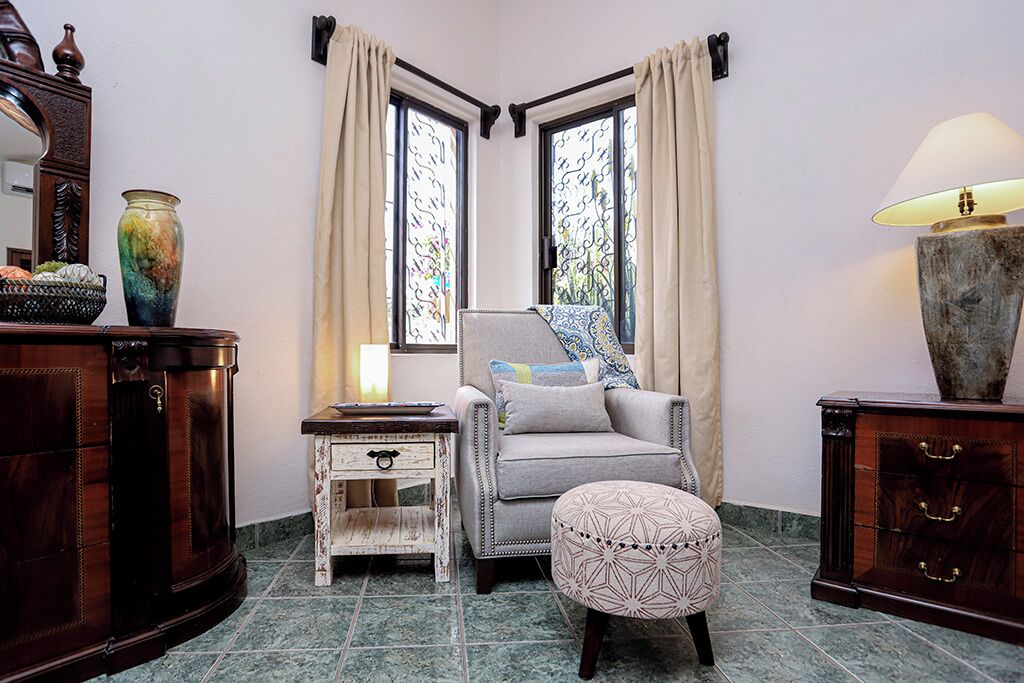 Habitación de esquina con una silla, reposapiés y una mesita de noche con lámpara. Se observan dos ventanas con cortinas, un espejo y un jarrón decorativo.