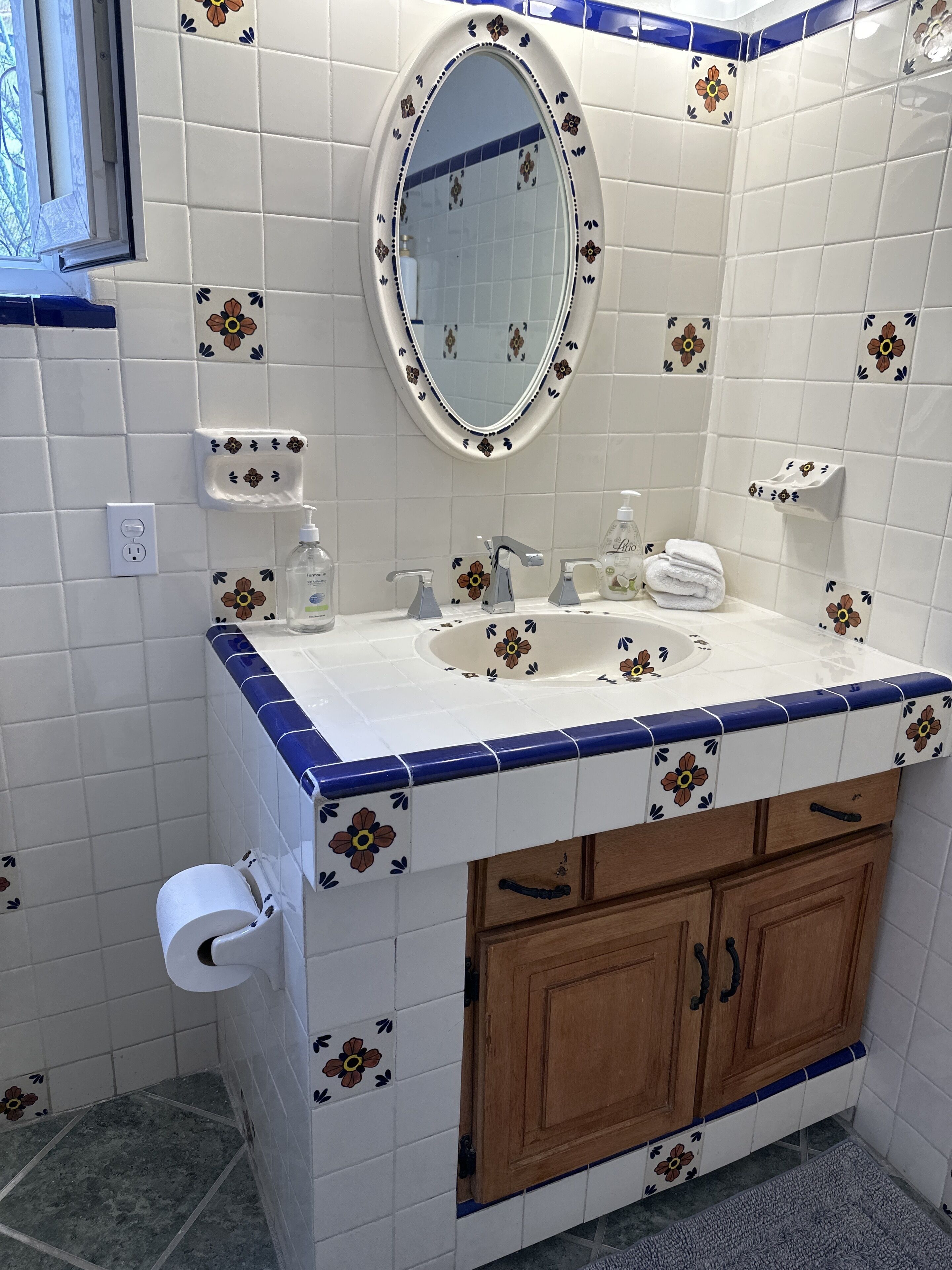 Et badeværelse med en hvid vask, blå flisearbejdsbordplade, træskabe, et spejl og en toiletpapirholder.