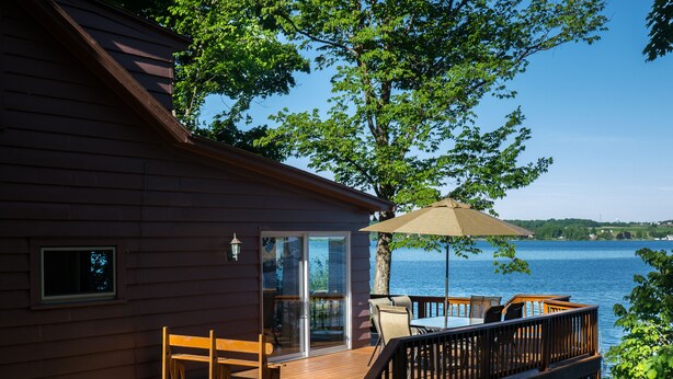 Eine Holzhütte mit einer Terrasse mit Blick auf einen See, ausgestattet mit Sitzgelegenheiten im Freien und einer Glastür, die zur Terrasse führt.