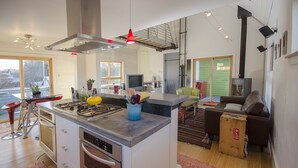 A modern kitchen with a central island, built-in oven, and stainless steel appliances. There is a dining area with a green sofa and a small table.